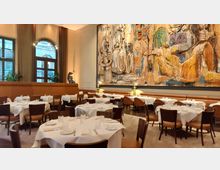 Elegantes Restaurant mit gedeckten Tischen, weißen Tischdecken und braunen Stühlen. Eine große, bunte Wandmalerei dominiert den Raum, daneben hohe Fenster mit Vorhängen und braunen Ledersitzbänken im Hintergrund.