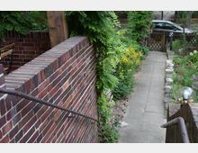 Ein gepflasterter Gartenweg führt an einer Backsteinmauer entlang, die mit einem Metallhandlauf versehen ist. Auf der rechten Seite des Weges befinden sich blühende Pflanzen und Sträucher; im Hintergrund sind ein Holztor und ein parkendes Auto erkennbar.