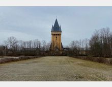 Turm einer Backsteinkirche mit spitzem Dach und kleinen Fenstern, flankiert von kahlen Bäumen, im Hintergrund niedrige Gebäude; im Vordergrund eine gepflasterte Freifläche.