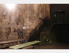 Großer, industrieller Innenraum mit einer abgenutzten, teilweise mit Graffiti bedeckten Betonwand. Eine Metalltreppe führt zu einer erhöhten Plattform, daneben stehen einfache Holzbänke und Tische.