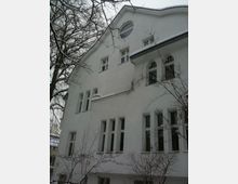 Wohnhaus mit weiß verputzter Fassade, hohen Fenstern im klassischen Stil und einem geschwungenen Giebel. Im Vordergrund sind kahle Äste von Bäumen im winterlichen Umfeld zu sehen.