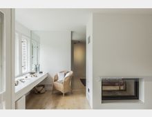 Heller, minimalistischer Wohnraum mit Holzboden, einem weißen Einbauschreibtisch unter mehreren Fenstern, einem Rattansessel mit Kissen und einem modernen, eingebauten Kamin in einer weißen Trennwand.