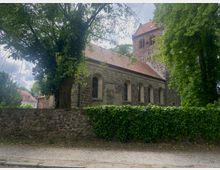 Historische Kirche aus Feldstein mit rotem Ziegeldach und Rundbogenfenstern, umgeben von Bäumen und einer Steinmauer, im Hintergrund weitere Gebäude sichtbar.
