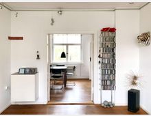 Minimalistisch eingerichtetes Wohnzimmer mit Holzboden und weiß gestrichenen Wänden. Im Vordergrund ein CD-Regal, eine kleine Skulptur und Dekorationsgegenstände; im Hintergrund ein angrenzender Essbereich mit modernem Tisch und Stühlen vor einem Fenster.