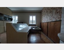 Ein retro-industrieller Raum mit einer großen Schaltkonsole und mehreren eingebauten Monitoren an der Wand. Im Hintergrund ein Fenster mit vergittertem Blick nach draußen sowie eine Tapete mit Blumenmuster und Holzpaneelen an der rechten Wand.