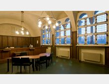 Geräumiger Sitzungssaal mit gewölbter Decke, großen Fenstern mit kleinen Scheiben und Holzrahmen sowie einer hölzernen Wandverkleidung. Zentral stehen ein langer Holztisch und mehrere Stühle; im Hintergrund befindet sich ein erhöhtes Podium.