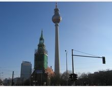 Der Berliner Fernsehturm ragt hoch in den Himmel, daneben die Marienkirche mit grünem Turmhelm, teilweise von einem Baugerüst und einer Abdeckung umgeben. Im Hintergrund sind moderne Gebäude und klare blaue Himmel zu sehen, im Vordergrund befinden sich Ampeln und Straßenlaternen.