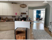 Helle, offene Wohnung mit moderner Küche im Vordergrund, ausgestattet mit cremefarbenen Schränken, Spüle und Holzdetails. Im Hintergrund ein Essbereich mit großen, bodentiefen Fenstern, blauer Wandfarbe und einem Holzboden in Fischgrätmuster.