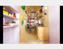 Ein kompakter Raum mit Wänden und Möbeln aus Holz, eingerichtet als Arbeits- und Wohnbereich. Links und rechts befinden sich Regale mit bunten Dekorationsgegenständen und Büchern, in der Mitte ein Bürostuhl vor einem kleinen Schreibtisch, rechts eine minimalistische Küchenzeile mit Spüle.