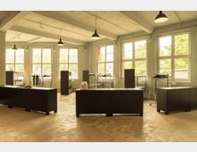 Ein minimalistisch eingerichteter Raum mit großen Fensterfronten, die viel Tageslicht hereinlassen. Der Raum enthält dunkle, moderne Theken und Regale sowie Kleiderständer, während die freiliegende Decke mit Hängelampen beleuchtet wird.