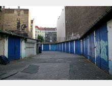 Ein Hinterhof mit einer Reihe von blauen Garagentoren, flankiert von hohen, teils verfallenen Backstein- und verputzten Wänden. Im Hintergrund sind mehrstöckige Wohnhäuser mit bunten Fassaden und roten Dächern zu sehen.