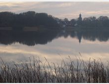Seeufer mit Schilf im Vordergrund, ein ruhiger See mit Spiegelung von Bäumen und Gebäuden in der Mitte, am Horizont eine kleine Kirche mit Turm, umgeben von Bäumen unter bewölktem Himmel.