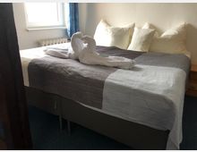 Ein schlicht eingerichtetes Schlafzimmer mit einem Doppelbett, das eine graue Tagesdecke und mehrere weiße Kissen hat. Auf dem Bett liegt ein dekorativ gefaltetes Handtuch in Form eines Schwans, im Hintergrund ein Fenster mit blauem Vorhang.