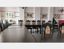 Ein Konferenzraum mit langen schwarzen Holztischen und reich verzierten Stühlen mit gepolsterten, gemusterten Lehnen. An den weißen Wänden hängen eingerahmte Bilder und Plakate, daneben sind ein Blumenstrauß und Bücher auf einer Kommode sichtbar.