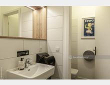 Kleines Badezimmer mit weiß gefliesten Wänden, einem Waschbecken mit Seifenspender und einem Dyson-Händetrockner. Im Hintergrund ist eine halb geöffnete Tür zu sehen, die zu einer Toilette führt; darüber hängt ein Poster mit der Aufschrift "Fewa".