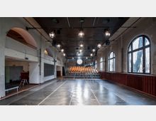 Raum mit Bühnen- und Aufführungscharakter, gekennzeichnet durch eine leere Tanzfläche im Vordergrund, Stuhlreihen auf einer Zuschauertribüne im Hintergrund und große Rundbogenfenster entlang der Wände. Die Decke ist mit mehreren schwenkbaren Scheinwerfern und Audioequipment ausgestattet.