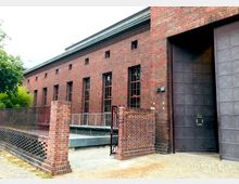 Backsteingebäude mit hohen Fenstern und Stahlrahmen; auffällig eine große, verzierte Metalltür und ein gemauerter Eingangsbereich, umgeben von einem Geländer aus Metall. Links ist ein Baum zu sehen, der den Eingang teilweise einrahmt.