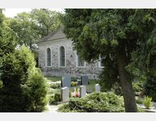 Steinbau mit Satteldach und schmalen, rundbogigen Fenstern, umgeben von einem Friedhof mit Grabsteinen, blühenden Blumen und dichter Bepflanzung; im Vordergrund Bäume und Sträucher.