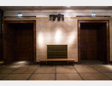 Innenansicht eines Gebäudeflurs mit hellen Steinwänden und einem Metallgitter in der Mitte, darunter eine schlichte Holzbank. Links und rechts befinden sich zwei große Türen mit Holzrahmen, umgeben von Marmorverkleidungen.