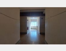 Ein einfacher Flur mit cremefarbenen Wänden und dunklem, glänzendem Bodenbelag. Am Ende des Flurs befindet sich ein Fenster, das Tageslicht hereinlässt, sowie ein Feuerlöscher, der an der linken Wand angebracht ist.