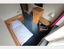 Kleines Hotelzimmer mit Einzelbett, blauem Teppichboden und Möbeln aus hellem Holz, darunter ein Kleiderschrank, ein Schreibtisch mit Mülleimer und ein Stuhl. Ein Fenster mit rosa Vorhängen und eine Tischlampe sind ebenfalls sichtbar.