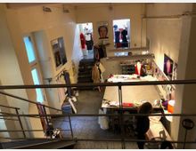 Ein heller, mehrstöckiger Arbeitsraum mit einer zentralen Werkbank, die mit Stoffen und Werkzeugen bedeckt ist. Links führt eine Treppe nach oben, Kleider hängen an der Wand, und im Hintergrund sind Schaufensterpuppen und Nähutensilien sichtbar.