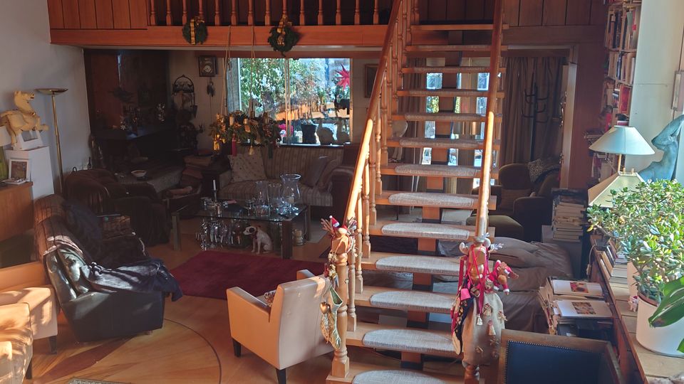 Wohnzimmer mit holzverkleideter Decke und Galerie im oberen Bereich, die über eine hölzerne Treppe mit geschmücktem Geländer erreichbar ist. Das Zimmer ist mit Teppichen, Polstermöbeln, einem Bücherregal und Dekorationen ausgestattet, und große Fenster lassen Tageslicht herein.