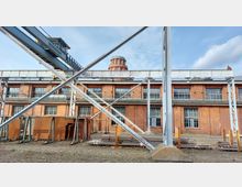 Industriegebäude aus rotem Backstein mit großen Fenstern und einem runden Turm im Hintergrund. Im Vordergrund sind Metallträger, Gerüste und gestapelte Baumaterialien, umgeben von einem unbefestigten Boden zu sehen.
