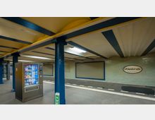 U-Bahn-Station mit gelb-blauer Decke und blau gestrichenen Stützsäulen. Im Vordergrund steht ein Snack- und Getränkeautomat, im Hintergrund sind geflieste Wände mit dem Stationsschild „Uhlandstraße Kurfürstendamm“ sichtbar.
