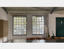 Industrieartiger Innenraum mit hoher Betondecke und sichtbaren Trägern, zwei großen Rasterfenstern und einem langen Tisch mit Stühlen im Vordergrund. Rechts befindet sich eine dunkle Tür sowie hängende Glühbirnen und einige Pflanzen.