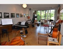 Proberaum mit Holzboden, ausgestattet mit Musikinstrumenten wie Klavier, Cello, Pulten mit Partituren und Stühlen. Große Fenster im Hintergrund bieten einen Blick ins Grüne, und an den Wänden hängen gerahmte Fotos.