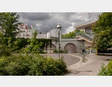 Straßenszene mit einer Grünanlage im Vordergrund, einem grauen Fußgängerweg und einem überdachten Treppenaufgang rechts. Im Hintergrund ist eine von Graffiti bedeckte Wand mit Rundbögen zu sehen, daneben mehrere Altbauwohnungen mit weißen Fassaden und roten Dächern unter bewölktem Himmel.