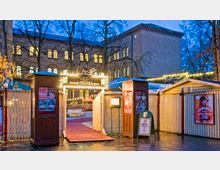 Eingang zu einem Veranstaltungsort mit der Aufschrift „Bar jeder Vernunft“ in Berlin, umgeben von beleuchteten Holzpavillons und einem roten Teppich. Im Hintergrund befindet sich ein Backsteingebäude mit erleuchteten Fenstern, umgeben von kahlen Bäumen.