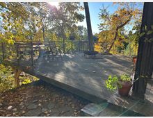 Holzterrasse inmitten von herbstlichem Laubwald, mit einfachem Geländer und Gartenmöbeln, darunter ein Tisch und Stühle. Im Hintergrund ist ein Feuerkorb zu sehen, während Sonnenlicht durch die bunten Blätter fällt.