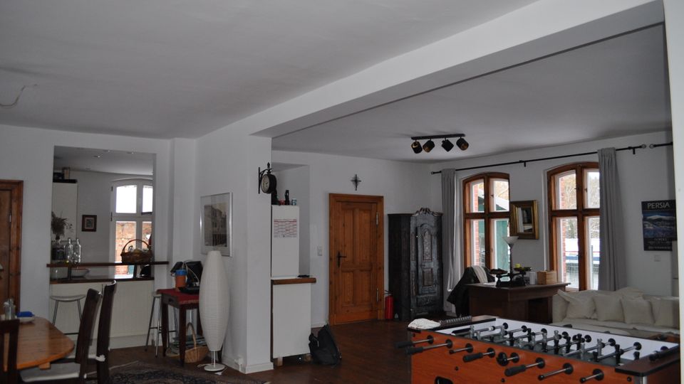 Wohnraum mit offenen Bereichen, weißen Wänden und Holzböden. Der Raum umfasst mehrere Fenstersätze mit Rundbögen, eine Küchenzeile, einen Esstisch, einen Kickertisch und einen Schrank aus dunklem Holz.