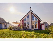 Ein rotes, modernes Ferienhaus mit Satteldach und großen Fenstern, umgeben von einer Rasenfläche und anderen ähnlich gestalteten Häusern. Im Vordergrund befinden sich Gartenmöbel, ein Sonnenschirm und ein Außenkamin.