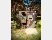 Camoufliertes Metalltor mit der Aufschrift „Sperrzone - Restricted Area“, umgeben von dichter Vegetation und Bäumen in einem Waldgebiet.