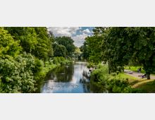 Ein Fluss, umgeben von dichtem Grün und großen Bäumen, fließt durch einen Park mit Spazierwegen und Sitzbänken. Schwäne schwimmen im Wasser, und im Hintergrund ist eine kleine Metallbrücke zu sehen.