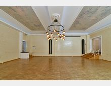 Ein großer, heller Raum mit Parkettboden und Wänden aus gelblichen Ziegelsteinen. Die Decke ist mit dekorativen Paneelen verziert, und in der Mitte hängt ein großer Kronleuchter mit mehreren Glühbirnen. Rechts führt eine kleine Treppe zu einem erhöhten Bereich mit Vorhangabtrennung.