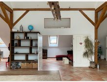 Wohnraum mit offenem Dachstuhl und sichtbaren Holzbalken, ausgestattet mit einem Holzregal, einer Zimmerpflanze und Fliesenboden. Im Hintergrund ein angrenzender Raum mit Dachfenstern, rotem Sofa und kleinem Couchtisch.