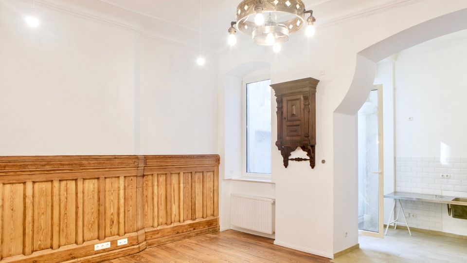 Heller, weitgehend leerer Innenraum mit weißen Wänden, Holzdielenboden und einer langen Holzvertäfelung entlang der linken Wand. An der Decke hängt ein Kronleuchter, neben einem Fenster ist ein dunkler Wandschrank montiert, und ein Rundbogen führt in einen gefliesten Nebenraum mit Glastür.