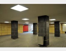 Unterirdischer Durchgangsbereich mit quadratischen dunklen Säulen, gelben Wandplatten und einer roten Wand im Hintergrund. Die Decke ist mit rechteckigen Leuchtflächen ausgestattet, und im Hintergrund führen Treppen nach oben.