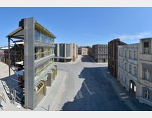 Straßenszene in einem Film- oder Studiogelände, mit Gebäudefassaden, die verschiedene Architekturstile nachahmen, darunter moderne Glas- und Stahlstrukturen sowie historische Fassaden. Die Umgebung ist leer, mit sandigem Untergrund und einem klaren blauen Himmel im Hintergrund.