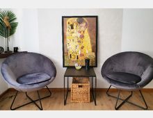 Ein kleiner Sitzbereich mit zwei runden, grauen Sesseln, einem schwarzen Beistelltisch dazwischen und einem gerahmten Bild („Der Kuss“ von Gustav Klimt) an der Wand. Auf dem Tisch stehen Dekorationsobjekte, darunter eine kleine Vase, und darunter befindet sich ein geflochtener Korb.