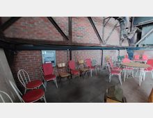 Ein rustikaler Raum mit Ziegelwänden, Metallträgern und alten, weißen Metallstühlen mit roten und holzfarbenen Sitzflächen. Im Raum stehen mehrere Tische und Stühle, die wie in einem industriellen oder historischen Gebäude arrangiert sind.