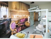 Moderne Zahnarztpraxis mit einem Behandlungsstuhl im Vordergrund und Arbeitsbereich mit Computer sowie Instrumenten. Eine auffällige Wandgestaltung zeigt ein historisches, opulent eingerichtetes Zimmer mit lila Polstermöbeln und goldenen Verzierungen.