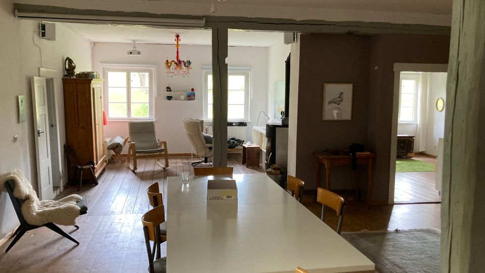 Offener Wohnbereich mit Holzboden, weißen Wänden und Balkendecke. Im Vordergrund steht ein großer weißer Esstisch, im Hintergrund sind Fenster, ein Holzschrank und Sitzmöbel erkennbar, die Zimmer durch breite Durchgänge verbunden.