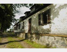 Ein historisches, weiß gestrichenes Gebäude mit bröckelnder Fassade und Fenstern mit hölzernen Fensterläden sowie Gitterstäben. Im Vordergrund führt ein schmaler, von Bäumen gesäumter Weg entlang des Gebäudes, umgeben von Gras und Schatten.