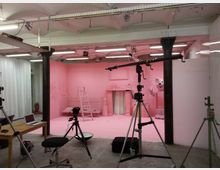 Ein Fotostudio oder Filmset mit einem rosa dekorierten Raum im Hintergrund, ausgestattet mit einer Leiter, einem Kamin und einem großformatigen Figurenkopf einer weißen Maus. Im Vordergrund befinden sich Kamerastative, Beleuchtungsgeräte und ein Tisch mit einem Laptop.