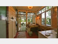 Ein schmaler, rustikaler Innenraum mit Holzwänden und großen Fenstern auf der rechten Seite, die auf einen Garten blicken. Im Hintergrund führt eine verglaste Tür zu einer überdachten Terrasse mit Blick ins Grüne.